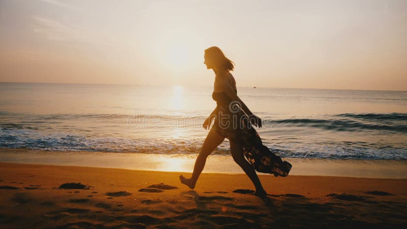 O meio feliz bonito envelheceu caminhadas e corridas da mulher ao longo da praia dourada épico do mar do por do sol que sorri na