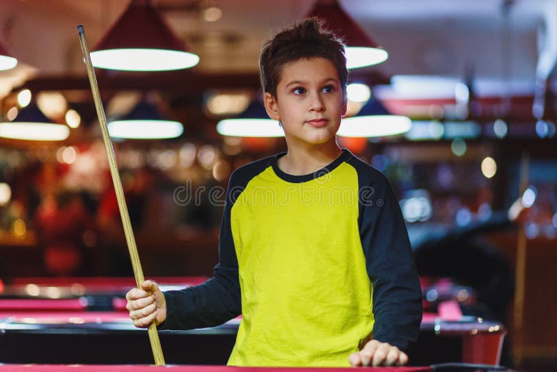 O menino bonito na camisa amarela de t joga o bilhar ou a associação no clube A criança aprende jogar a sinuca Menino com sugestã fotografia de stock royalty free O menino bonito na camisa amarela de t joga o bilhar ou a associação no clube A criança aprende jogar a sinuca Menino com sugestã fotografia de stock royalty free