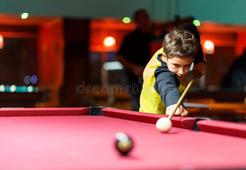 O menino bonito na camisa amarela de t joga o bilhar ou a associação no clube A criança aprende jogar a sinuca Menino com sugestã imagem de stock O menino bonito na camisa amarela de t joga o bilhar ou a associação no clube A criança aprende jogar a sinuca Menino com sugestã imagem de stock