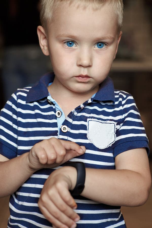 O menino louro pequeno bonito com olhos azuis indica ao perseguidor digital em sua expressão séria do pulso, emoções fortes da ap imagem de stock O menino louro pequeno bonito com olhos azuis indica ao perseguidor digital em sua expressão séria do pulso, emoções fortes da ap imagem de stock