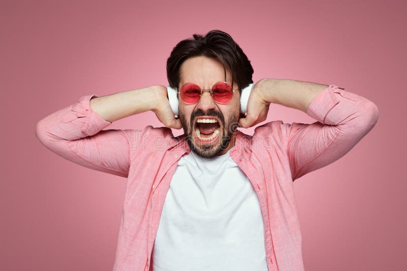O menino novo emocional mantiver as mãos nos fones de ouvido brancos, gritos quando música de escuta com o volume alto, isolado s imagem de stock royalty free O menino novo emocional mantiver as mãos nos fones de ouvido brancos, gritos quando música de escuta com o volume alto, isolado s imagem de stock royalty free