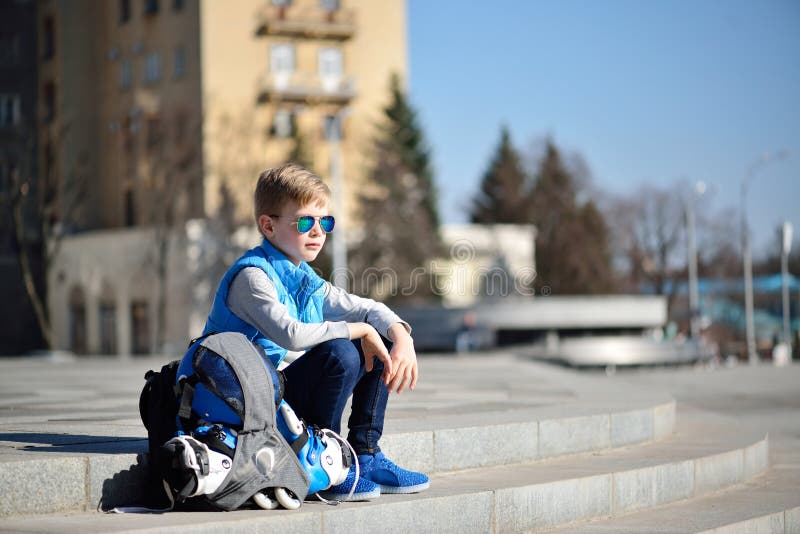 O menino novo que sentam-se perto da parede cinzenta com os patins de rolo inline e toda a proteção no patim exterior estacionam imagem de stock royalty free O menino novo que sentam-se perto da parede cinzenta com os patins de rolo inline e toda a proteção no patim exterior estacionam imagem de stock royalty free
