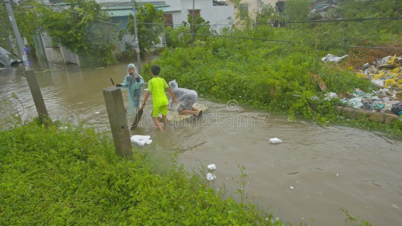 O menino tem a natação do divertimento na caminhada dos amigos da prancha ao longo da rua inundada