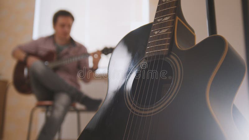 O músico atrativo novo compõe a música na guitarra e os jogos, o outro instrumento musical no primeiro plano, borrado foto de stock O músico atrativo novo compõe a música na guitarra e os jogos, o outro instrumento musical no primeiro plano, borrado foto de stock