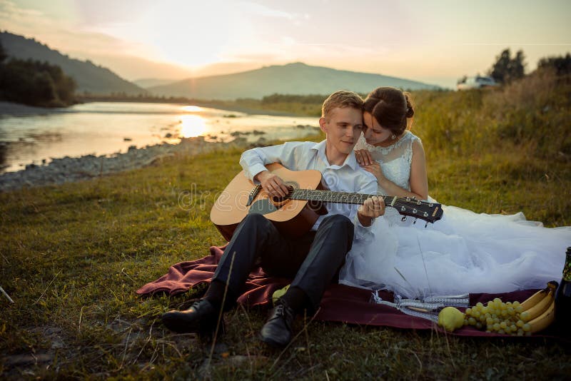 O noivo atrativo joga a guitarra e sua noiva encantador aprecia-a durante seu piquenique no banco de rio durante foto de stock O noivo atrativo joga a guitarra e sua noiva encantador aprecia-a durante seu piquenique no banco de rio durante foto de stock