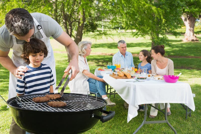 O pai e o filho no assado grelham com a família que tem o almoço no parque imagens de stock royalty free O pai e o filho no assado grelham com a família que tem o almoço no parque imagens de stock royalty free