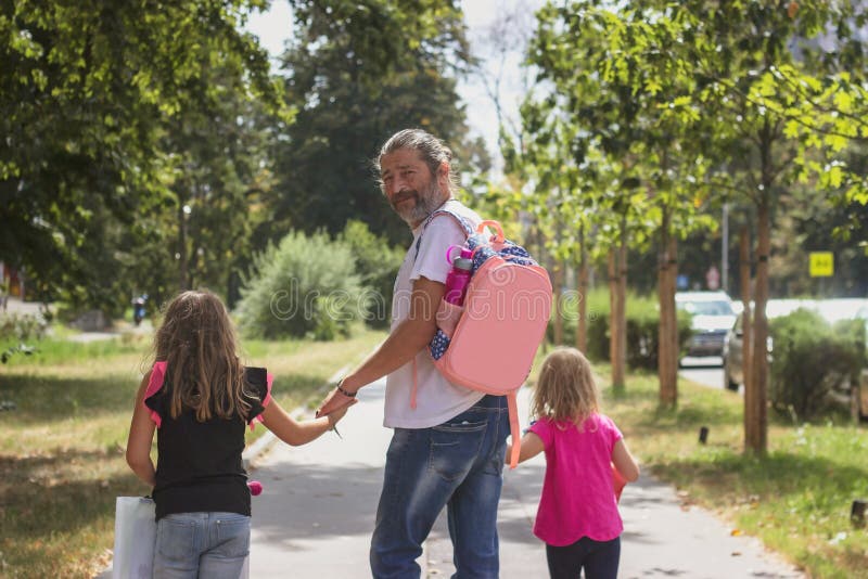 O pai está a levar as suas filhas para a escola, a vestir a bolsa das crianças nas costas imagens de stock O pai está a levar as suas filhas para a escola, a vestir a bolsa das crianças nas costas imagens de stock