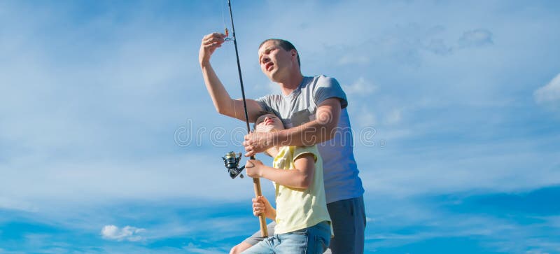 O pai mostra seu filho no cais, como estabelecer uma vara de pesca para travar peixes, contra o céu azul, close-up imagem de stock O pai mostra seu filho no cais, como estabelecer uma vara de pesca para travar peixes, contra o céu azul, close-up imagem de stock