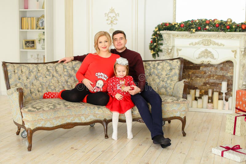 O pai novo e a mãe grávida que sentam-se com a filha pequena no sofá próximo decoraram a chaminé foto de stock O pai novo e a mãe grávida que sentam-se com a filha pequena no sofá próximo decoraram a chaminé foto de stock