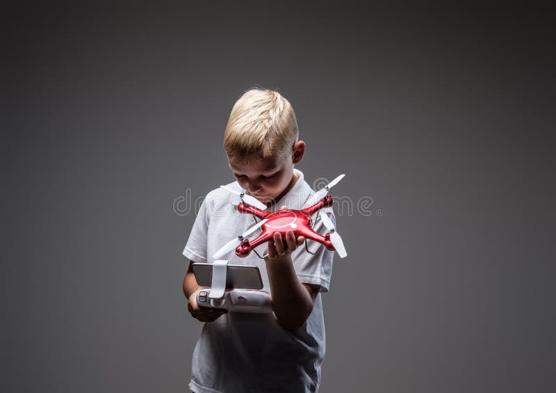 O pugilista considerável do rapaz pequeno com o cabelo louro vestido em um t-shirt branco guarda um telecontrole do quadcopter e imagens de stock O pugilista considerável do rapaz pequeno com o cabelo louro vestido em um t-shirt branco guarda um telecontrole do quadcopter e imagens de stock