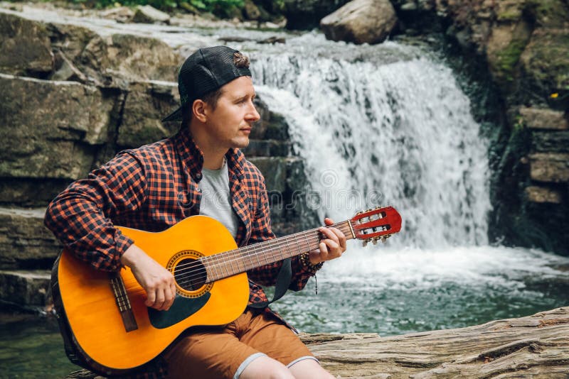 O retrato de um homem joga uma guitarra que senta-se em um tronco de uma árvore contra uma cachoeira Espaço para sua mensagem de fotografia de stock O retrato de um homem joga uma guitarra que senta-se em um tronco de uma árvore contra uma cachoeira Espaço para sua mensagem de fotografia de stock
