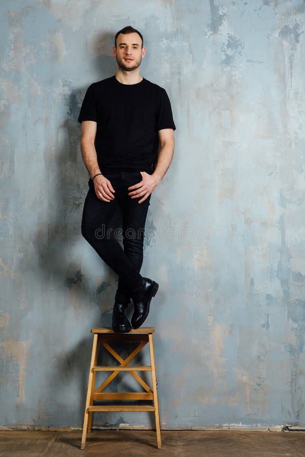 O retrato de um homem novo em um t-shirt preto inclinou-se contra uma parede do cinza do vintage Copie o espaço imagens de stock O retrato de um homem novo em um t-shirt preto inclinou-se contra uma parede do cinza do vintage Copie o espaço imagens de stock