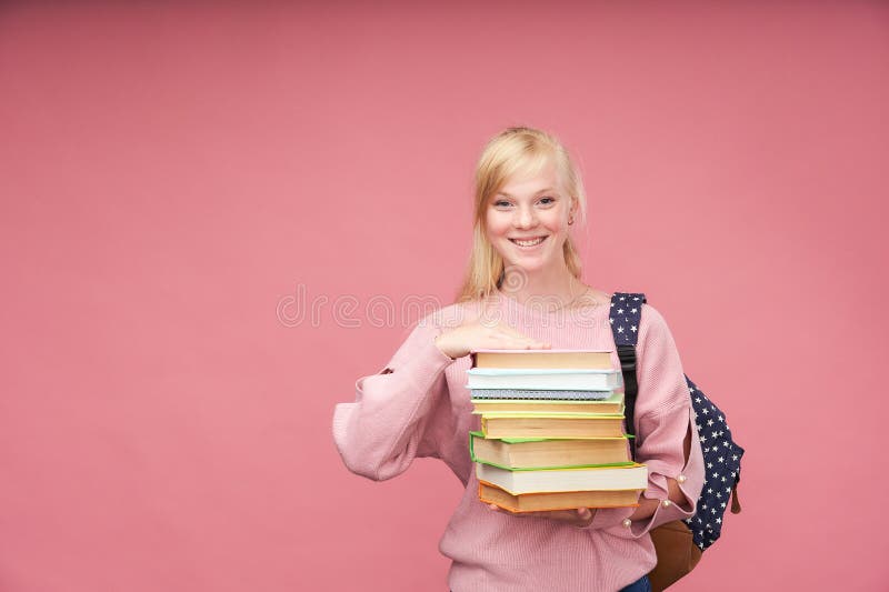 O retrato de uma estudante bonita com uma trouxa e de uma pilha de livros em suas mãos está sorrindo no fundo cor-de-rosa imagens de stock O retrato de uma estudante bonita com uma trouxa e de uma pilha de livros em suas mãos está sorrindo no fundo cor-de-rosa imagens de stock