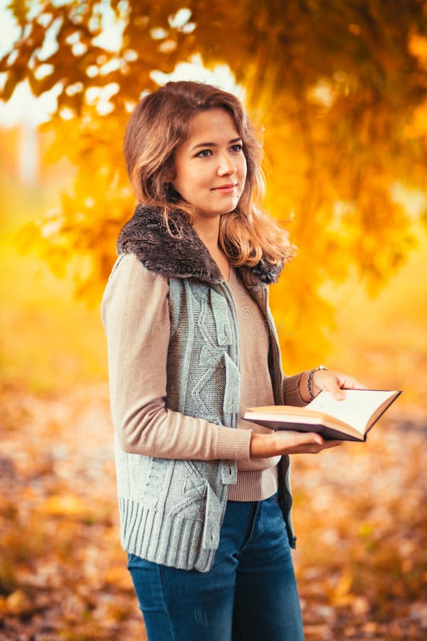 O retrato de uma moça na veste da pele e o livro de leitura no outono do fundo estacionam fotografia de stock O retrato de uma moça na veste da pele e o livro de leitura no outono do fundo estacionam fotografia de stock