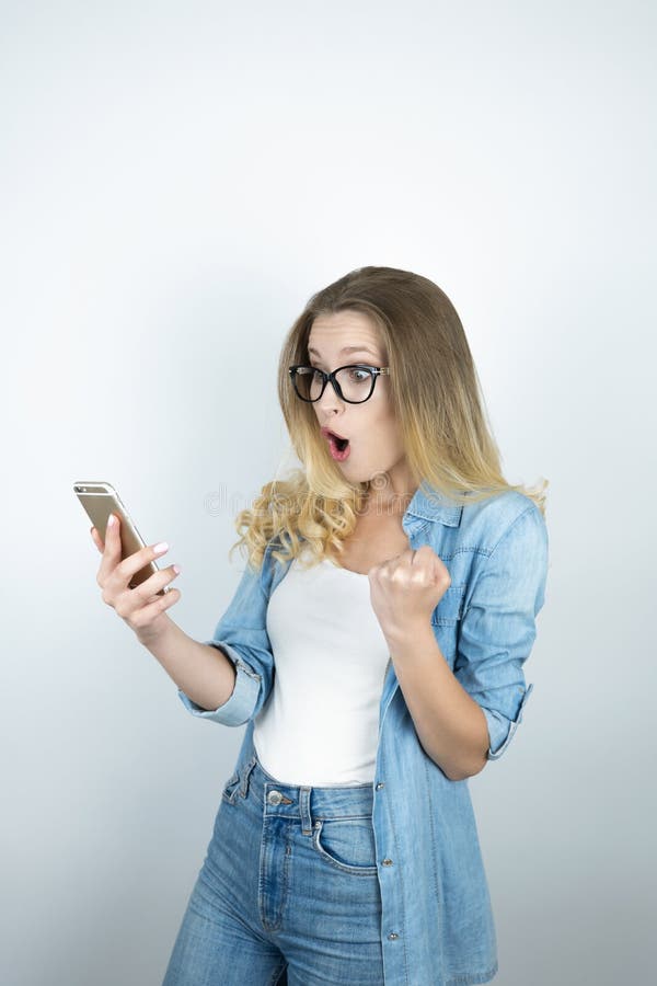 O smartphone surpreendido louro novo bonito da terra arrendada da mulher olha o fim feliz acima do fundo branco imagens de stock O smartphone surpreendido louro novo bonito da terra arrendada da mulher olha o fim feliz acima do fundo branco imagens de stock