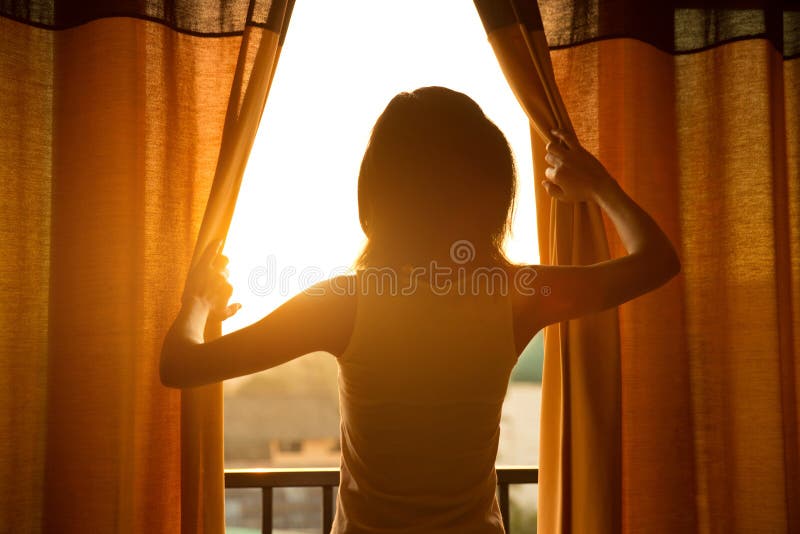 O suporte da mulher na cortina aberta da sala considera o nascer do sol imagens de stock royalty free O suporte da mulher na cortina aberta da sala considera o nascer do sol imagens de stock royalty free