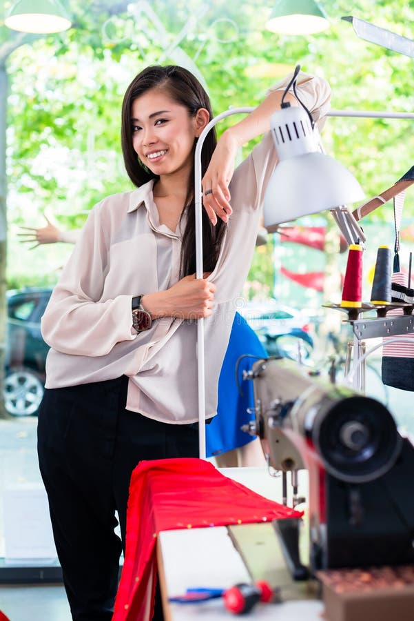 O tainlor ou o desenhador de moda asiático inclinam-se na mesa da costura imagem de stock O tainlor ou o desenhador de moda asiático inclinam-se na mesa da costura imagem de stock