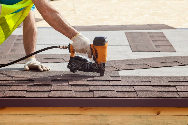 O trabalhador da construção que põe as telhas do telhado de asfalto com prego atira em uma casa de quadro nova O trabalhador da construção que põe as telhas do telhado de asfalto com prego atira em uma casa de quadro nova