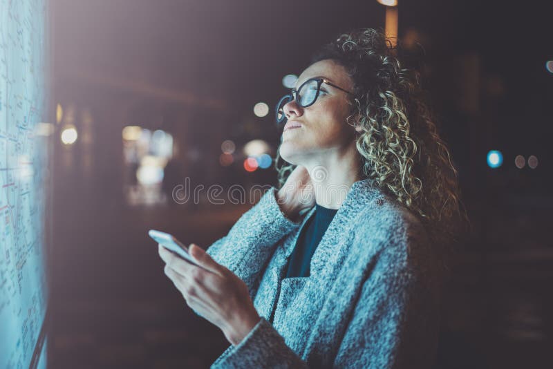 O turista encantador da mulher elegante com os vidros dos olhos que guardam o telefone celular entrega ao olhar o mapa da cidade imagem de stock royalty free O turista encantador da mulher elegante com os vidros dos olhos que guardam o telefone celular entrega ao olhar o mapa da cidade imagem de stock royalty free