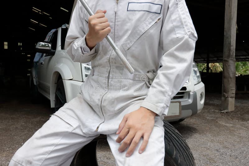 O ângulo largo disparou do mecânico profissional na chave e no pneu guardando uniformes no fundo da garagem do reparo Conceito do imagens de stock royalty free O ângulo largo disparou do mecânico profissional na chave e no pneu guardando uniformes no fundo da garagem do reparo Conceito do imagens de stock royalty free