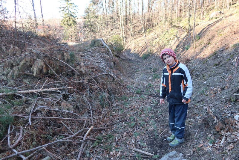 O întreagă figură de băiat pe un drum forestier într-un loc izolat. băiatul își înclină capul pe o parte și se uită în afară împuș. întreaga figură a unui băiat autist în haine de iarnă pe un drum forestier la locul unde pădurea a fost tăiată. există o grămadă de ramuri pe sol. restul pădurii de conifere este la orizont. băiatul are capul înclinat în fotografie și se uită spre stânga spre cameră. se pare că se distrează.