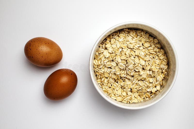 Oats in a Bowl and Two Raw Brown Eggs Aside, Top View on White