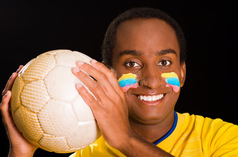 A obscuridade do Headshot descascou o homem que veste a camisa amarela do futebol na frente do fundo preto, pintura facial da ban imagens de stock royalty free A obscuridade do Headshot descascou o homem que veste a camisa amarela do futebol na frente do fundo preto, pintura facial da ban imagens de stock royalty free