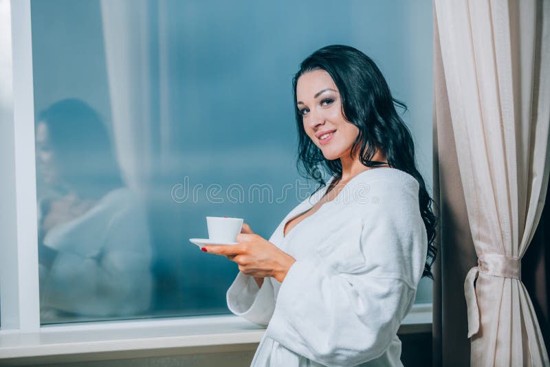 Obtention chaude avec du café frais Belle jeune femme en café potable de peignoir blanc et regard par une fenêtre photographie stock libre de droits Obtention chaude avec du café frais Belle jeune femme en café potable de peignoir blanc et regard par une fenêtre photographie stock libre de droits
