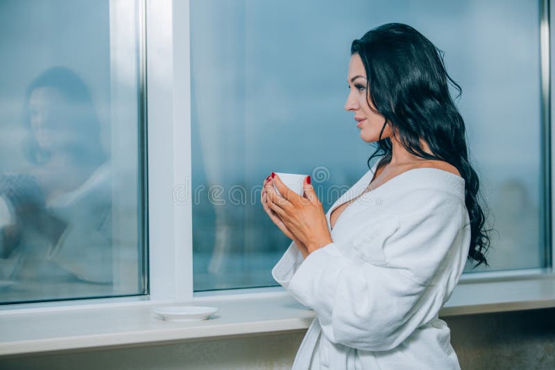 Obtention chaude avec du café frais Belle jeune femme en café potable de peignoir blanc et regard par une fenêtre image stock Obtention chaude avec du café frais Belle jeune femme en café potable de peignoir blanc et regard par une fenêtre image stock