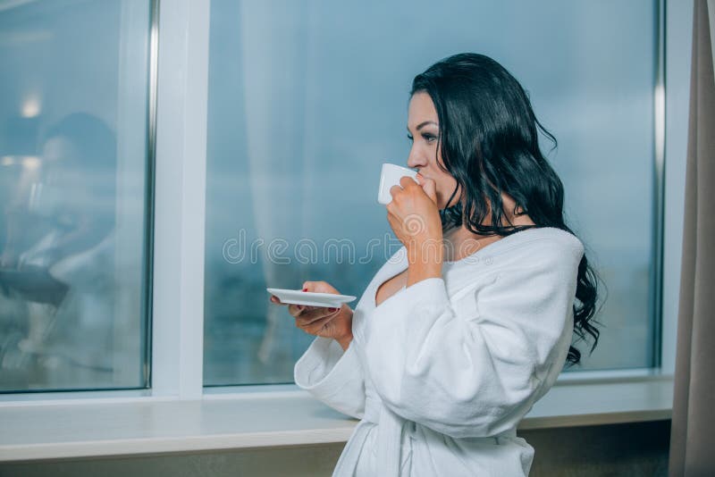 Obtention chaude avec du café frais Belle jeune femme en café potable de peignoir blanc et regard par une fenêtre image libre de droits Obtention chaude avec du café frais Belle jeune femme en café potable de peignoir blanc et regard par une fenêtre image libre de droits