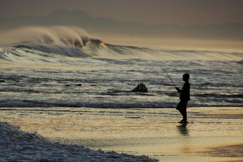 Silueta mladý chlapec, rybolov v ocean surf pri západe slnka.