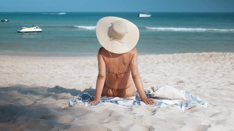 By ocean's edge woman sitting in bikini and hat on beach. Woman on beach salty breeze kissing skin rhythmic sound of