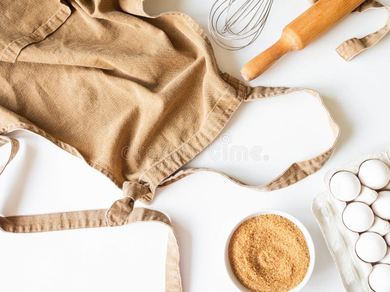 Ocher apron and various baking ingredients - eggs, sugar and kitchen utensils on white background. Top view