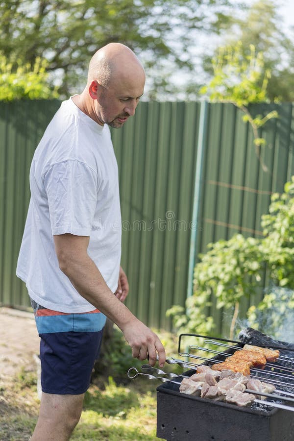 Ocio, comida, gente y concepto de los días de fiesta - hombre joven feliz que cocina la carne en parrilla de la barbacoa en el pa fotografía de archivo libre de regalías Ocio, comida, gente y concepto de los días de fiesta - hombre joven feliz que cocina la carne en parrilla de la barbacoa en el pa fotografía de archivo libre de regalías