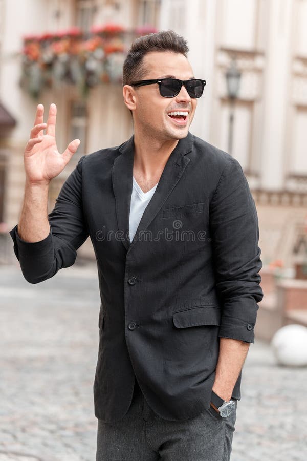 Ocio del aire libre Hombre joven y gafas de sol en la situación del traje en la calle de la ciudad que agita a la sonrisa del ami fotografía de archivo libre de regalías Ocio del aire libre Hombre joven y gafas de sol en la situación del traje en la calle de la ciudad que agita a la sonrisa del ami fotografía de archivo libre de regalías