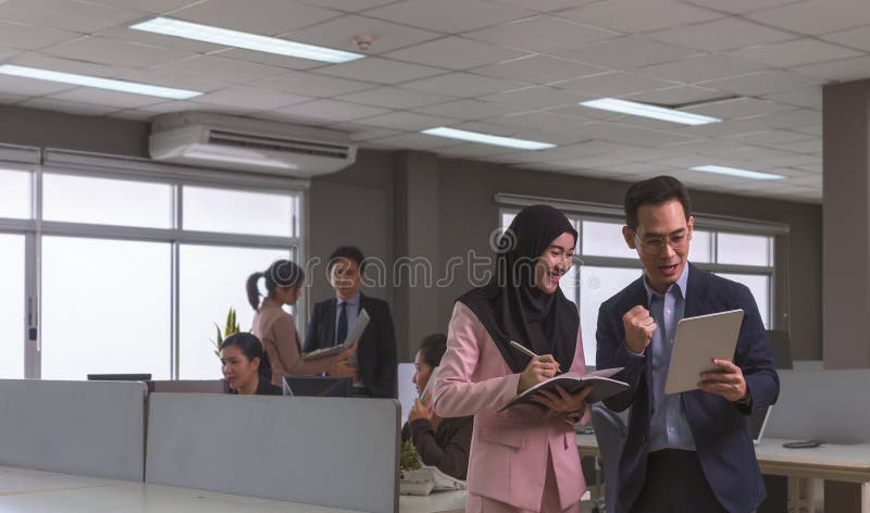 Office-anställd som tittar på digital Tablet PC medan han står på ett kontor med öppet utrymme med oskarpa kolleger som arbetar i. Lycklig affärsman i mörkblå dräkt med digital tablett som arbetar med muslimska kvinnor som bär hijab med anteckningar medan han står på kontor på öppet utrymme. arbetsplatskoncept för företagsgrupp