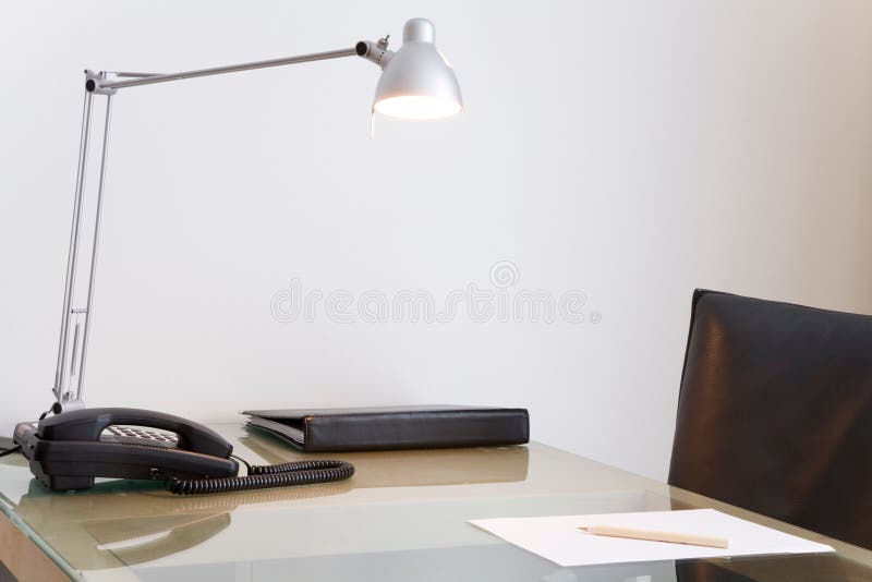 Desk with lamp and black leather swivel chair. White wall in the background. Desk with lamp and black leather swivel chair. White wall in the background.