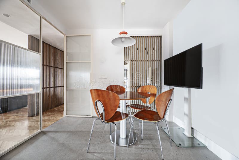 Office of a modern designed kitchen with a round glossy wooden table under a ceiling lamp, a flat tv on a stand, a glass partition