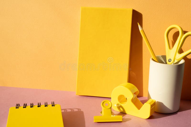 Office supply stationery. Yellow notebook, colored pencil, scissors, tape dispenser, clip on pink desk. orange wall background. workspace. studio shot