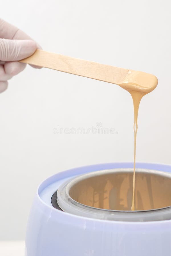 Wax heater with hot yellow wax on wooden spatula in human hand. Hot wax for depilation. Salon equipment. Wax heater with hot yellow wax on wooden spatula in human hand. Hot wax for depilation. Salon equipment