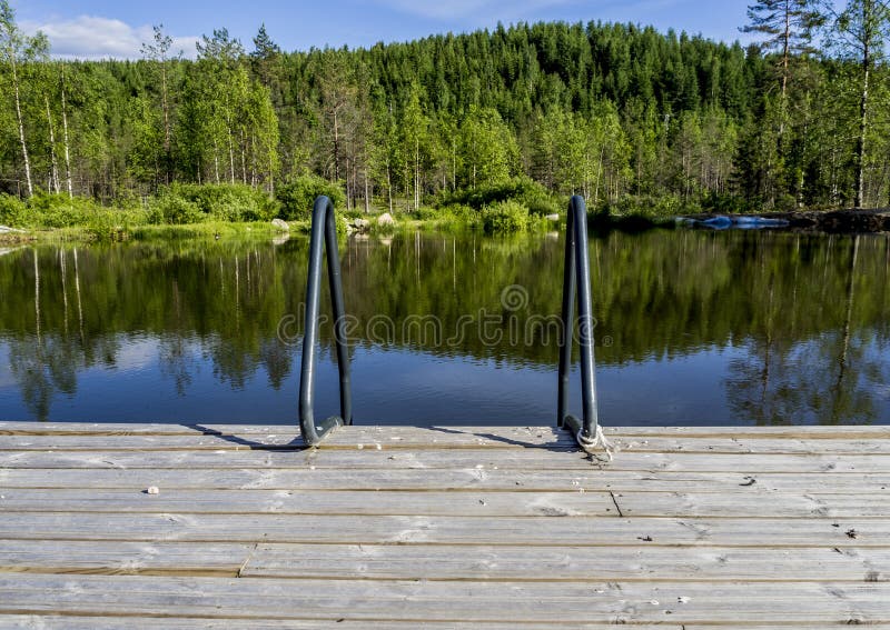 A pier with a ladder leading to the water, with a forest and hills in the background. Shot in NilsiÃ¤, Finland