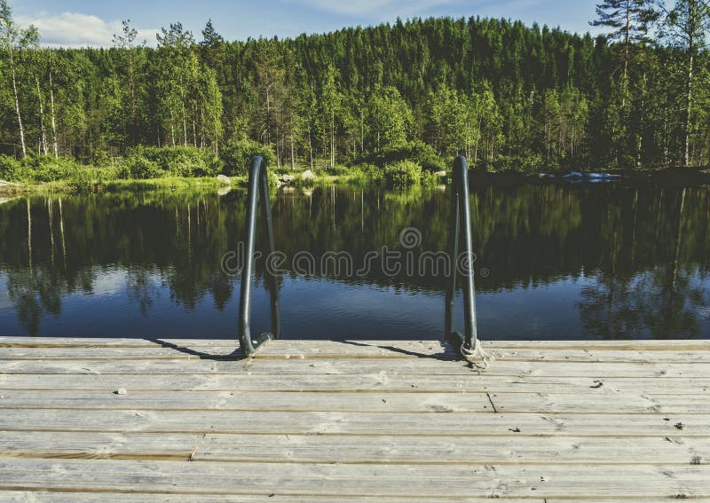 A pier with a ladder leading to the water, with a forest and hills in the background. Shot in NilsiÃ¤, Finland