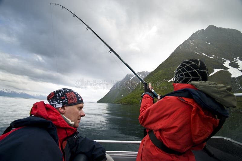 Father and son wearing red waterproof clothing fishing from boat, Norwegian fjord in background. Father and son wearing red waterproof clothing fishing from boat, Norwegian fjord in background.