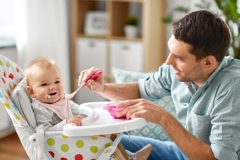 Ojciec karmi szczęśliwego dziecka w highchair w domu zdjęcie stock Ojciec karmi szczęśliwego dziecka w highchair w domu zdjęcie stock
