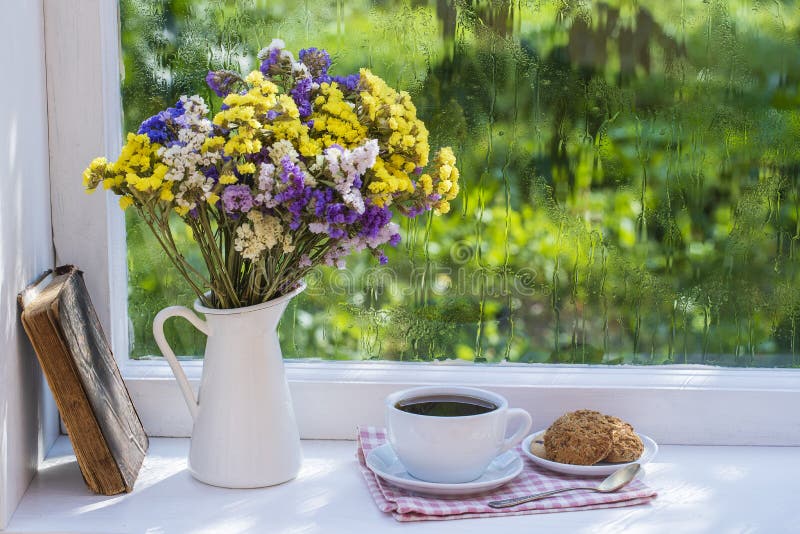 Old book, colorful bouquet of flowers and white cup of tea on background of window with raindrops at home at summer day