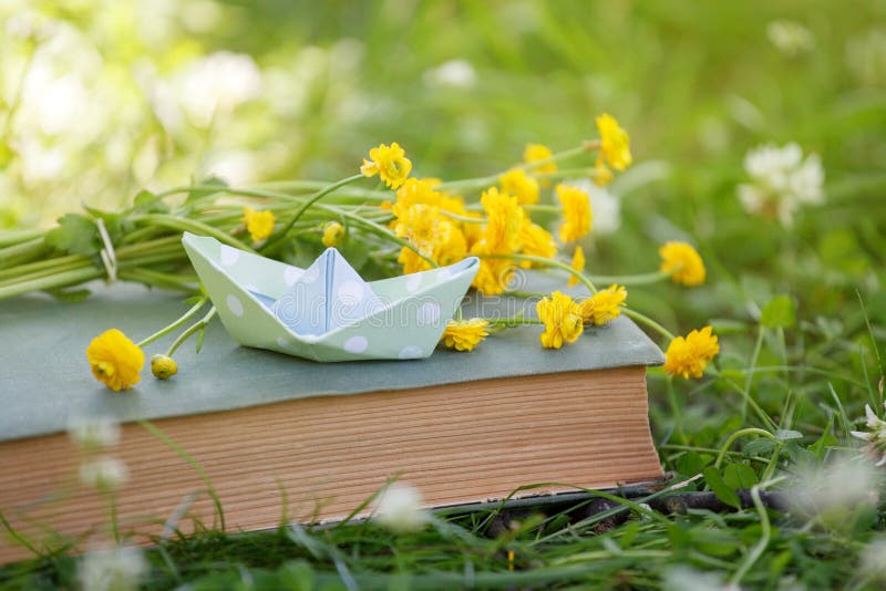 Old book, yellow flowers and papaer boat in green grass over yellow sun light. Magic book, time to dreaming, relax spring summer