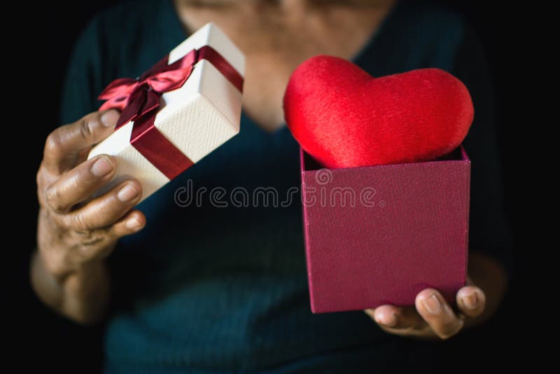 Old female hand holding gift box and red heart on black background stock photo Old female hand holding gift box and red heart on black background stock photo