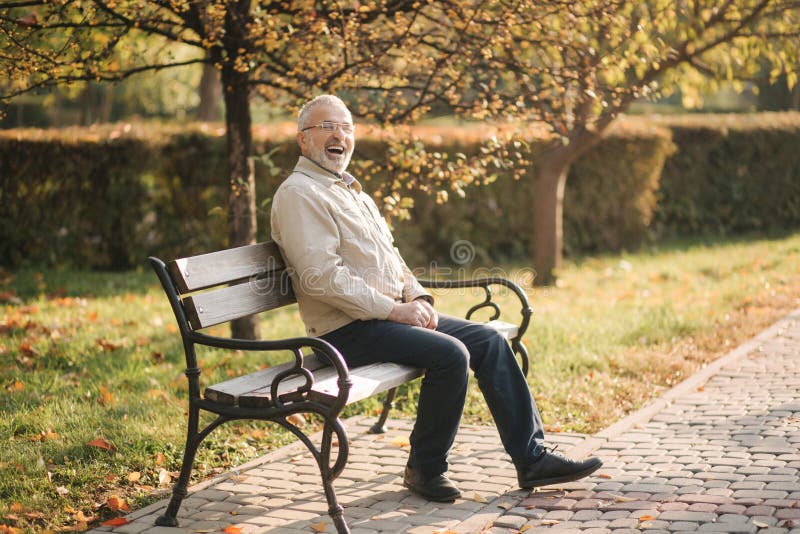 Old gray-haired man rest on the bench in autumn park. Elderly man smile