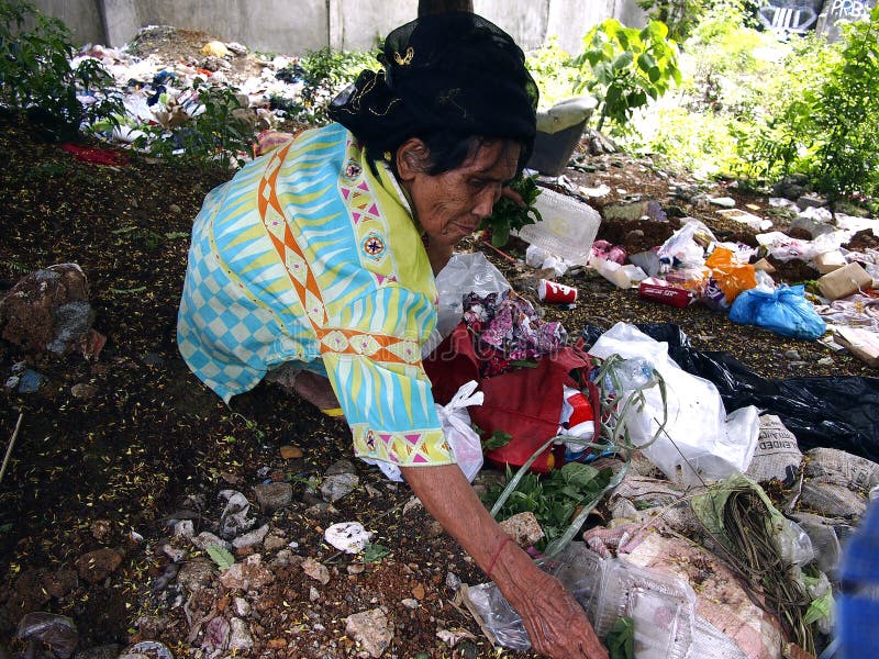 An old lady hunts or scavenges for recyclable materials in a pile of trash in an abandoned lot.