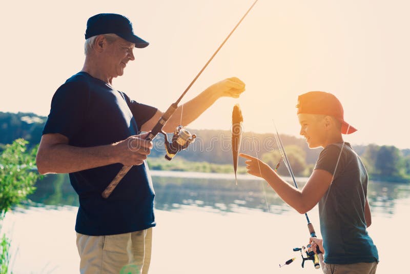 The old man and his grandson are standing on the river bank. The old man caught the fish and shows the catch to the boy